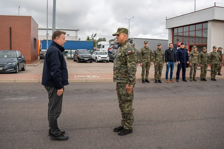 Mężczyzna w ciemnej kurtce rozmawia z żołnierzem w mundurze maskującym na parkingu przed budynkami, w tle stoją inni żołnierze i pojazdy.