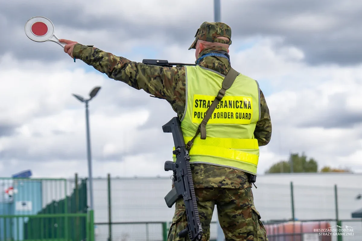 Funkcjonariusz polskiej Straży Granicznej w kamuflażu i żółtej kamizelce z napisem „STRAŻ GRANICZNA / POLISH BORDER GUARD” trzyma wyciągniętą ręką lizak do zatr