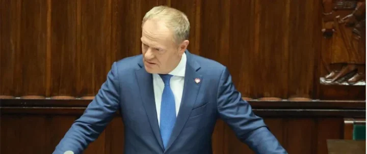 Mężczyzna w niebieskim garniturze i krawacie przemawia z mównicy w sali posiedzeń o drewnianych panelach.