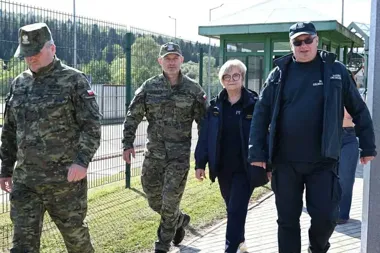 Cztery osoby, w tym dwóch żołnierzy w mundurach, kobieta w granatowej kurtce i mężczyzna w czapce z napisem "CELNICA", przechodzą obok ogrodzenia z budką strażn
