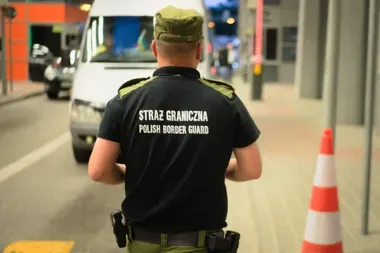 Funkcjonariusz Straży Granicznej stoi tyłem przy drodze, w tle widoczny biały samochód dostawczy oraz pachołek drogowy.