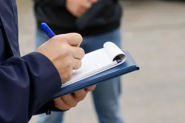 Funkcjonariusz w granatowej kurtce zapisuje coś długopisem na notesie, w tle stoi mężczyzna w czarnej kurtce i dżinsach.