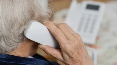 Starsza osoba trzyma słuchawkę białego telefonu stacjonarnego przy uchu.