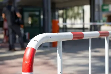 Czerwono-biały barierowy poręcz na zewnątrz, w tle rozmyta postać przechodnia i budynek.
